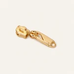 Assembled gold-tone detachable zipper pull tab and slider base shown on a warm beige background.