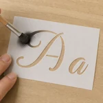 Hand using a brush to apply dark paint through an Aa stencil on wood; brush and paint are demonstration tools, not included.