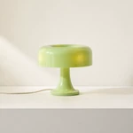 Light-green mushroom table lamp with rounded shade and pedestal base beside a cup on a console.