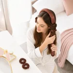 Mulher a enrolar o cabelo numa fita almofadada castanha junto de um espelho e elásticos