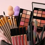 Nahaufnahme von Schminkpinseln, Lippenstiften, Paletten sowie pfirsichfarbenen und violetten Schwämmen am Kofferrand