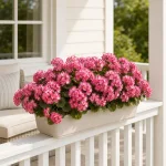 Pink artificial geranium flowers arranged in a white rectangular planter on a porch railing