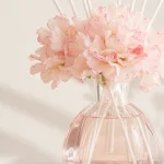 Nahaufnahme von rosa Blüten, hellen Stäbchen und dem Hals einer facettierten Diffuserflasche