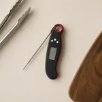 Draufsicht auf ein Lebensmittelthermometer mit geöffneter Sonde neben einer Zange und einem Holzbrett.