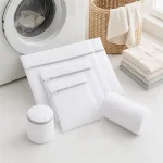 Weisse Wäschenetze in verschiedenen Formen neben Waschmaschine, Wäschekorb und gefalteten Handtüchern