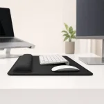 Black desk pad and wrist cushion beside a white keyboard and mouse between a laptop and monitor