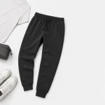 Jogger foncé posé à plat près d’une chemise blanche, d’une pochette grise et de baskets