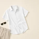 Camicia bianca a maniche corte abbottonata stesa accanto a pantaloni beige e occhiali da sole