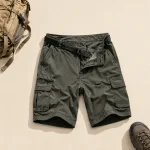 Flach ausgelegte olivgrüne Cargoshorts und ein schwarzer Gürtel neben Rucksack und Wanderschuh