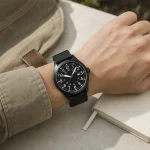 Black watch with a dark case worn on a wrist beside a notebook and khaki sleeve
