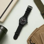 Black watch laid beside a notebook, separate green strap, bottle and tan bag
