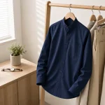Camisa azul marino con cuello mao en una percha de madera junto a pantalones claros y una mesita de noche