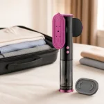 Vaporizzatore compatto per abiti accanto a una valigia aperta, vestiti piegati e un accessorio ovale.