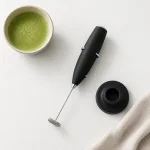 Montalatte nero e supporto rotondo appoggiati accanto a una ciotola di matcha