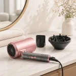 Pink hair dryer with nozzle and diffuser on a bathroom counter beside folded towels.