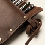 Gros plan sur une trousse à pinceaux marron montrant rivets, coutures de bord, lanière et manches de pinceaux