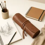 Trousse à pinceaux marron fermée par une lanière, près de carnets de croquis, d'un crayon et d'une tasse de pinceaux