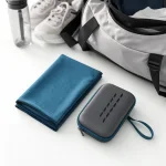 Toalla azul doblada y funda gris con cremallera junto a una bolsa de gimnasio, zapatos y botella de agua