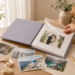 Hand lifting the clear overlay of a gray photo album beside loose prints on a table