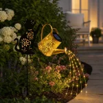 Gartenleuchte in Form einer Gießkanne auf einem Katzenstecker neben Blumen und einem Weg in der Dämmerung