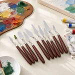 Palettmesser mit rotbraunen Griffen neben einer farbbedeckten Palette, Farbtuben und Leinwand