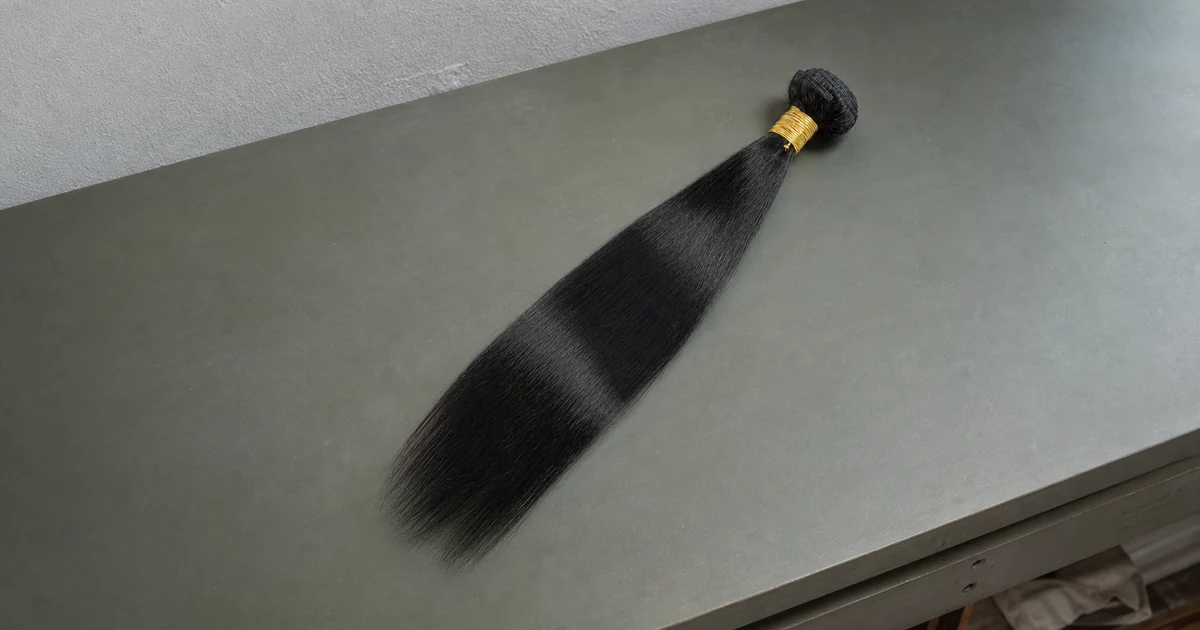 Ein glattes natur-schwarzes Haarbündel mit aufgerollter Tresse und goldfarbener Bindung auf einer graugrünen Arbeitsfläche.