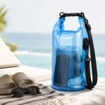 Bolsa azul enrollable sobre una toalla de playa junto a sandalias y gafas de sol, con el mar al fondo.