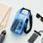 Vista superior de una bolsa azul enrollable junto a toalla, botella, gafas de sol y funda para teléfono.
