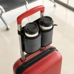 Zwei Take-away-Becher mit Deckel in einem dunklen Getränkehalter am ausgezogenen Griff eines roten Koffers