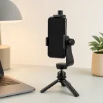 Soporte para teléfono mostrado en un pequeño trípode de escritorio junto a un portátil