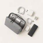 Bolsa tecnológica cinzenta aberta com cabos e adaptadores, junto de um telemóvel e estojo de auriculares