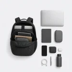 Mochila escura aberta mostrando compartimentos internos, com laptop, cadernos e pequenos acessórios dispostos ao lado
