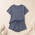 Blue-gray speckled pajama top and drawstring shorts laid flat beside slippers and a blanket