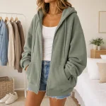 Frau mit offenem grünem Zip-Hoodie über weißem Tanktop und Jeansshorts drinnen