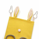 Détail supérieur d'un sac à foin jaune montrant des languettes en forme d'oreille, deux crochets et un pompon crème
