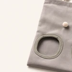 Gros plan sur un sac à foin gris montrant une ouverture d'alimentation bordée, la texture du tissu et un pompon crème