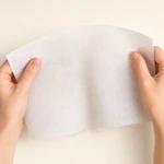 Two hands holding a single thin, flexible white nonwoven sheet against an ivory background