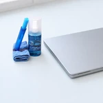 Frasco azul de limpeza, pano dobrado e escova dispostos ao lado de um laptop prateado fechado